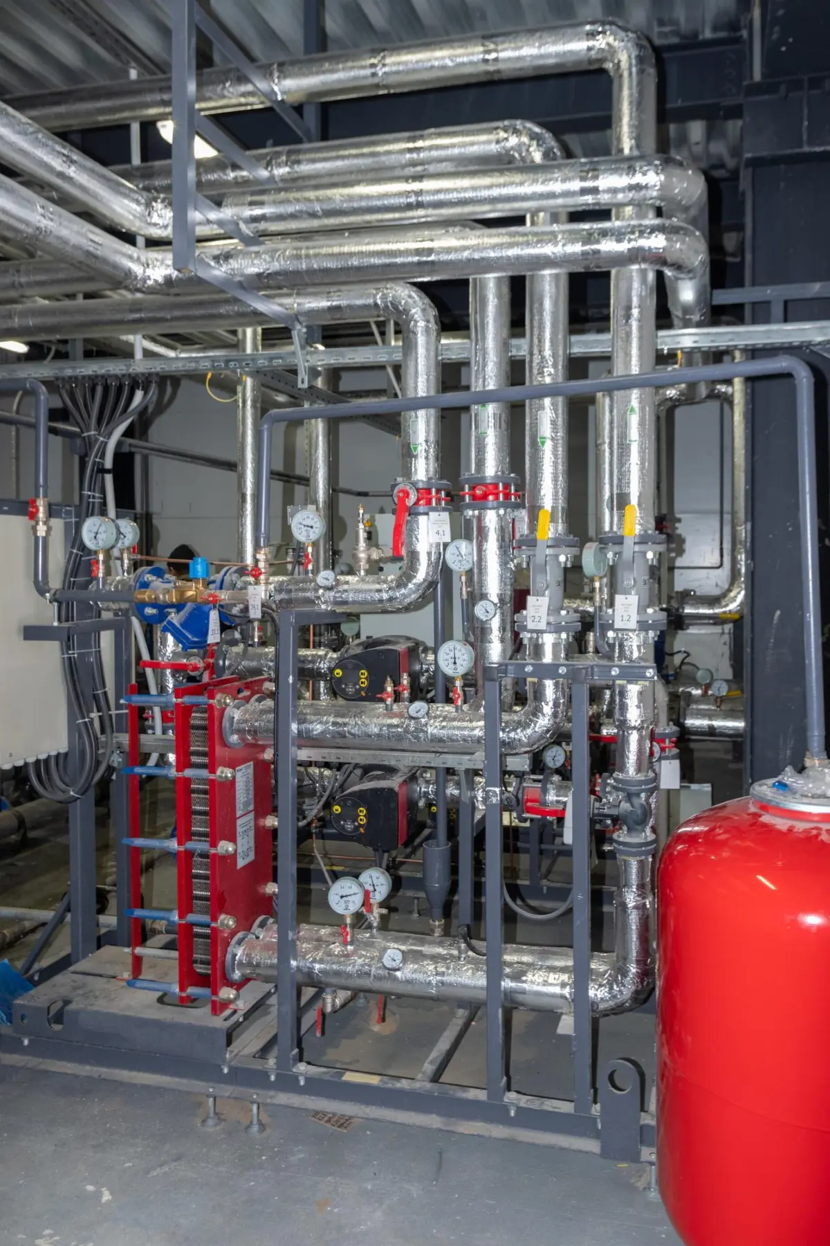 Technikraum mit Rohrleitungen, Pumpen, Manometern und rotem Ausdehnungsgefäß