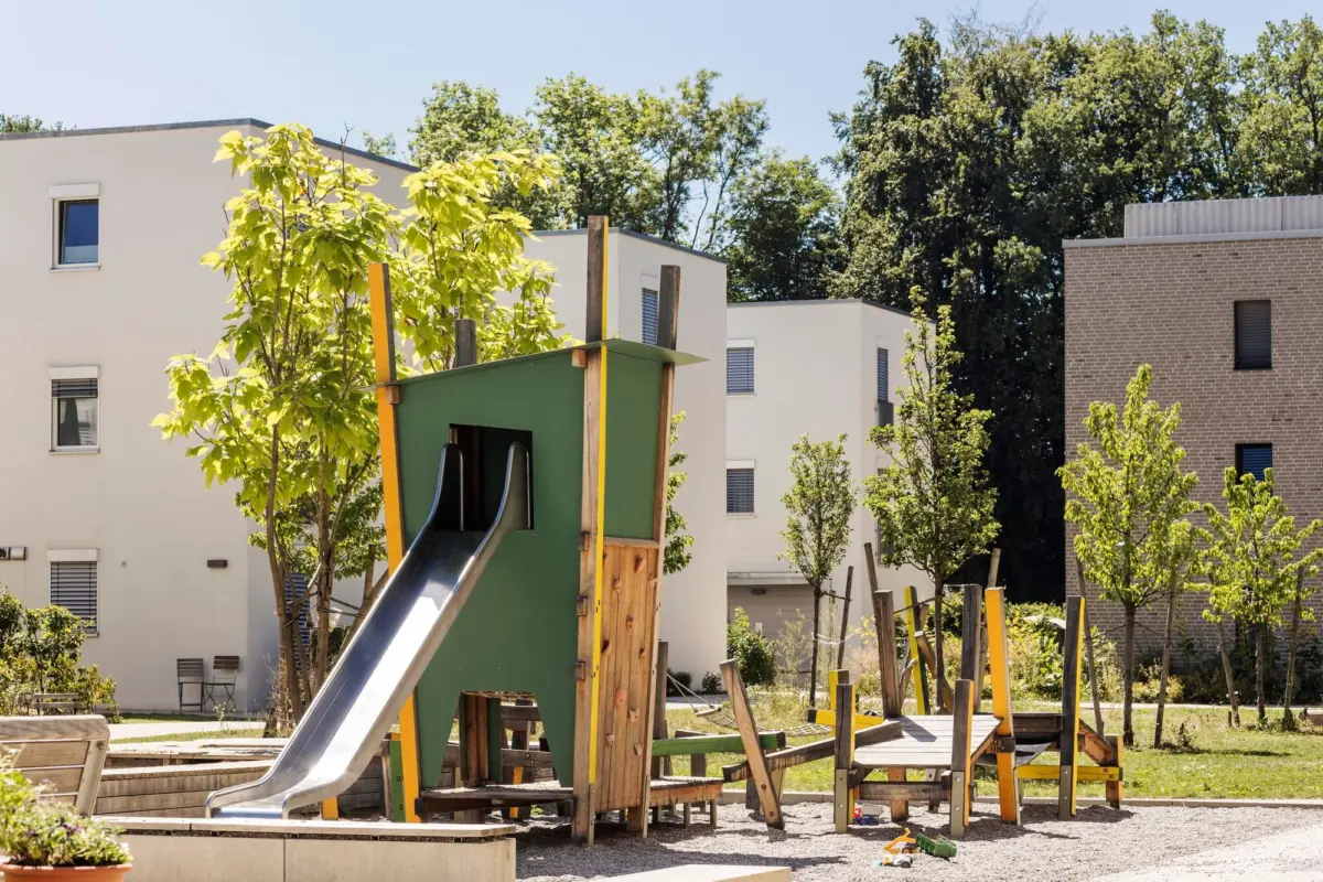 Spielplatz mit Holzturm und Rutsche zwischen hellen Gebäuden und Bäumen