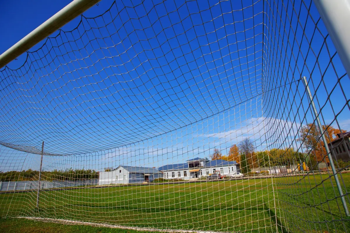 Blick durch das Netz eines Fußballtors auf einen Rasenplatz und ein Vereinsgebäude